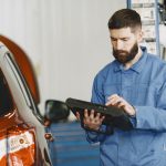 man in blue coveralls standing beside an orange car using an automotive diagnostic tool
