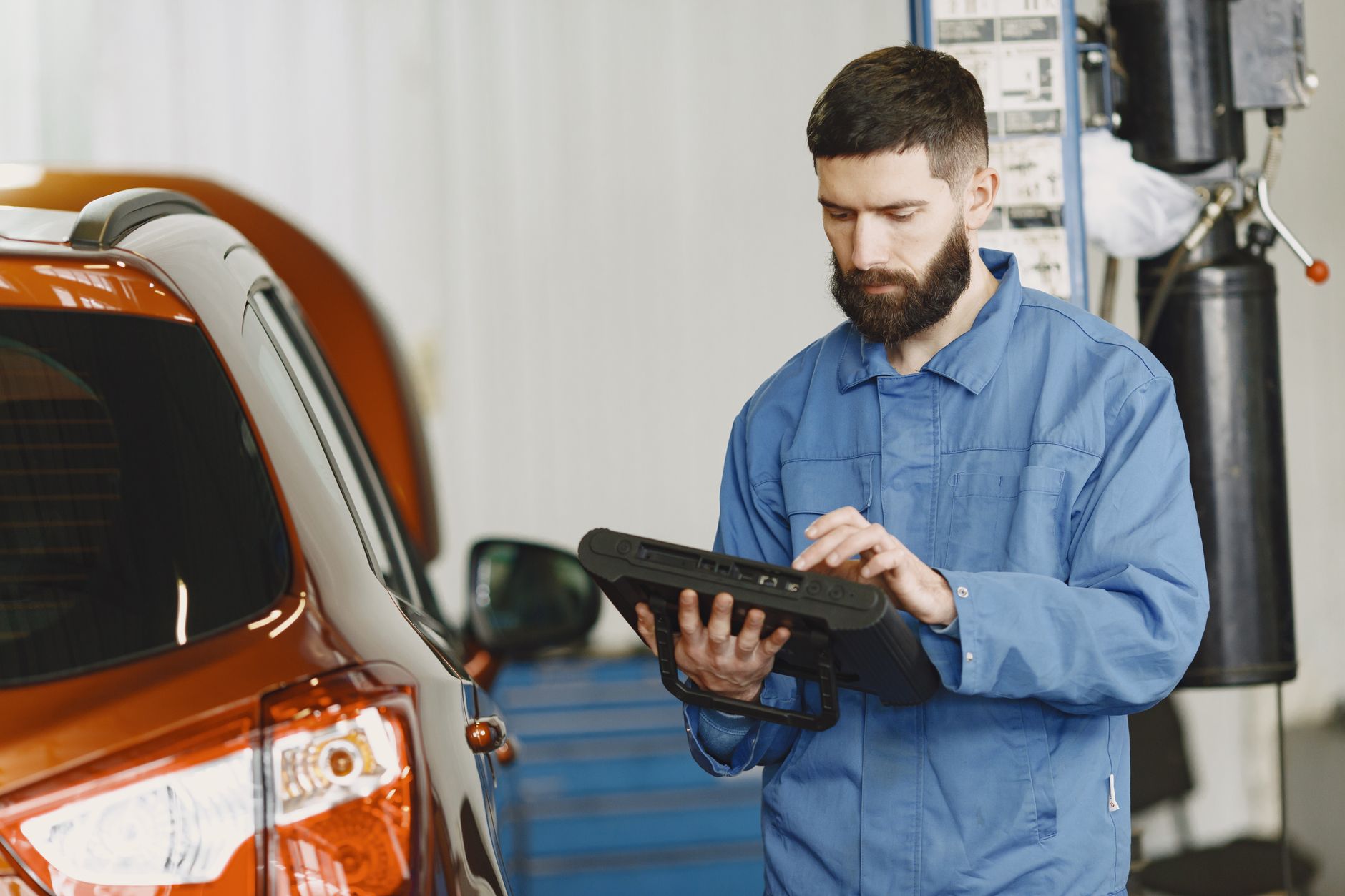man in blue coveralls standing beside an orange car using an automotive diagnostic tool